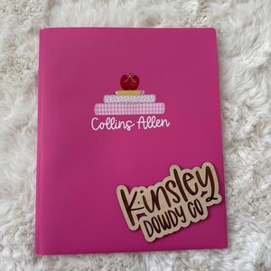 May include: A bright pink binder with a stack of books and an apple graphic, and the text "Collins Allen". A wooden sticker with the text "Kinsley Dowdy Co" is on the binder.