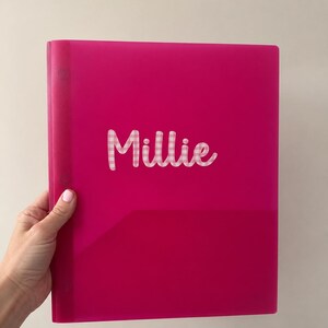 May include: A bright fuchsia plastic folder featuring the name Millie in white script lettering with a light pink and white gingham pattern fill. The folder has a vertical spine with visible circular fasteners along the left side edge.