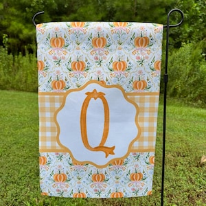 May include: A rectangular garden flag featuring a repeating pattern of orange pumpkins and small blue flowers on a white background. A central horizontal band displays a yellow gingham check pattern with a large, orange, stylized letter Q.