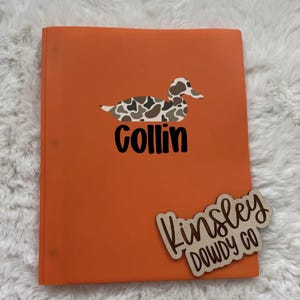 May include: An orange binder with a duck graphic and the name "Collin" in black. A wooden tag with the text "Kinsley DOWDY CO" is attached to the binder. The duck has a brown and white camouflage pattern.