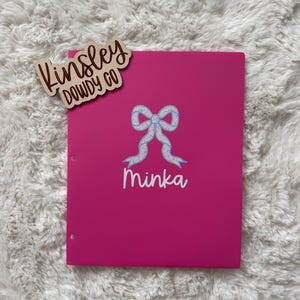 May include: A bright pink binder with a bow design and the name "Minka" in white script. A wooden tag with the text "Kinsley Dowdy Co" is attached to the binder. The binder is on a white, fluffy surface.