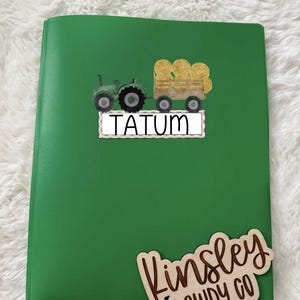 May include: A green notebook with a tractor and hay bale design. The name "TATUM" is printed on a white rectangle. A wooden cutout with the name "Kinsley DOWDY CO" is also visible.