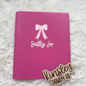 May include: A bright pink notebook with the name "Sally Jo" in white script and a gingham bow graphic. A wooden sign with the name "Kinsley Dowdy Co" is placed on the bottom right corner. The notebook is on a white fluffy surface.