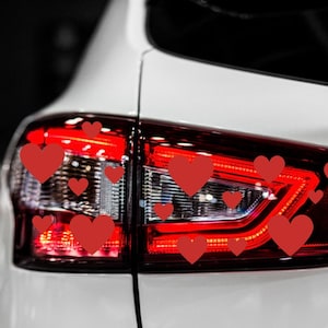 May include: A white car with a red taillight. The taillight is decorated with multiple red heart stickers.