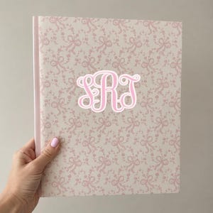 May include: A hardcover notebook featuring a light cream background with a repeating pattern of soft pink bows and floral sprigs. Centered on the front cover is a large, stylized monogram with the letters SRT in a bold, white-outlined pink font.