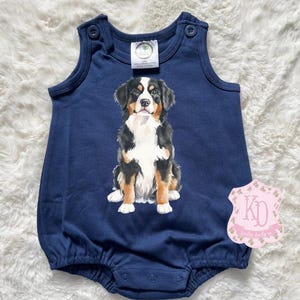 May include: A navy blue baby onesie with a white, black, and brown Bernese Mountain Dog illustration. The onesie has a sleeveless design and snaps at the bottom.