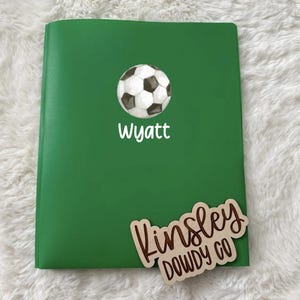 May include: A green notebook with a soccer ball graphic and the name "Wyatt" in white. A wooden tag with the name "Kinsley" and "DOWDY CO" is also present.