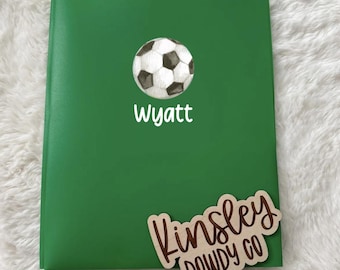 以下が含まれることがあります： 緑色のノート。サッカーボールのグラフィックと、白で「Wyatt」という名前が書かれています。「Kinsley」と「DOWDY CO」という名前の木製タグも付いています。
