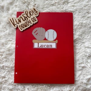 May include: A red binder with a baseball-themed design. The design includes a baseball glove, a baseball, and a baseball bat. The name "Lucan" is printed on the binder. A wooden cutout with the text "Kinsley Dowdy Co" is attached to the top.