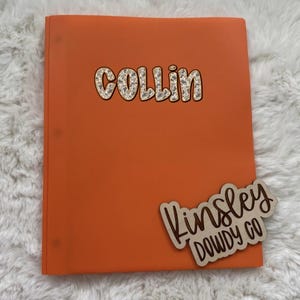 May include: An orange binder with the name "Collin" in a patterned font. A wooden name tag that says "Kinsley DOWDY CO" is attached to the binder.