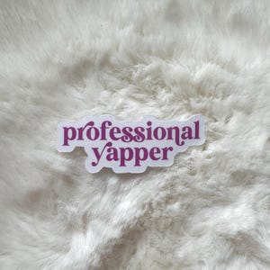 May include: A white sticker with the words "professional yapper" in a purple, cursive font. The sticker is placed on a fluffy, white surface.