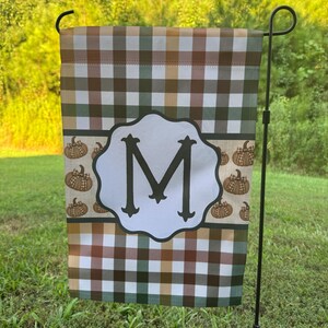 May include: A rectangular garden flag featuring a brown, green, and white plaid pattern. A central white scalloped emblem displays a large black letter M. The middle section includes a horizontal band decorated with small brown pumpkin illustrations.