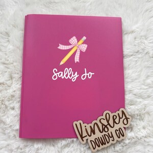 May include: A bright pink notebook featuring a yellow pencil graphic tied with a pink and white gingham bow. The name Sally Jo is printed in white script below the pencil. A wooden cutout sign with the brown text Kinsley Dowdy Co is present.
