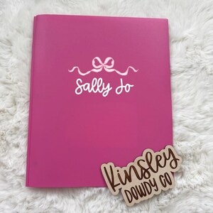 May include: A bright pink notebook with the name "Sally Jo" in white script, adorned with a bow design. A wooden tag with the words "Kinsley Dowdy Co" is placed on the bottom right corner.