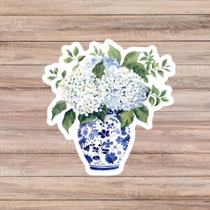 May include: A sticker featuring a watercolor illustration of a blue and white porcelain vase filled with white and light blue hydrangeas and green leaves. The vase has a floral pattern. The sticker is on a wooden background.