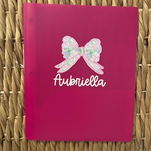 May include: A bright magenta plastic folder featuring a central graphic of a bow with a light pink, blue, and green floral pattern. Below the bow, the name Aubriella is printed in a white, cursive script font on the front cover.