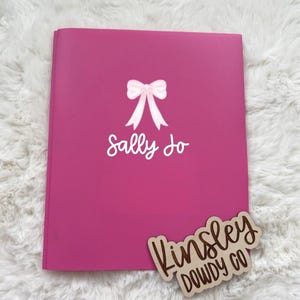 May include: A bright pink notebook with a white bow and the name "Sally Jo" printed on the cover. A wooden tag with the text "Kinsley Dowdy Co" is placed on the bottom right corner.
