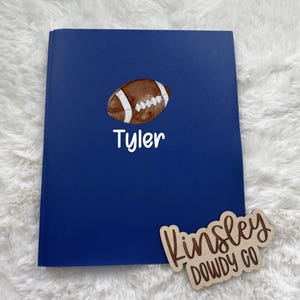 May include: A navy blue notebook with a watercolor football illustration and the name "Tyler" in white script. A wooden sign with the text "Kinsley DOWDY CO" is next to the notebook. The notebook is on a white fluffy surface.