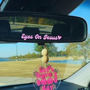 May include: A car rearview mirror with the pink text "Eyes On Jesus" above a decorative car charm. The charm is clear with the text "Let's have a good day" and smiley faces in pink, surrounded by floral accents.