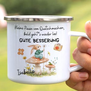 Könnte beinhalten: Weißer Emaille-Becher mit silbernem Rand, mit einer skurrilen Illustration eines Hasen auf einem Pilz, einem Pflaster und einem Erste-Hilfe-Kasten. Der Becher hat Text auf Deutsch und den Namen "Isabella".