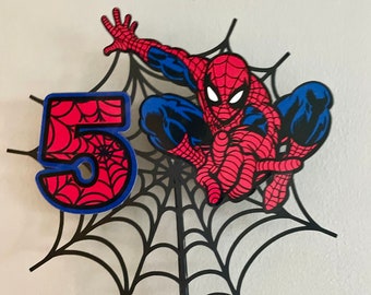 May include: A Spiderman-themed cake topper featuring the number 5 in red with a spiderweb design and blue trim. The Spiderman figure is in a dynamic pose, set against a black spiderweb background. The topper is attached to a wooden stick.