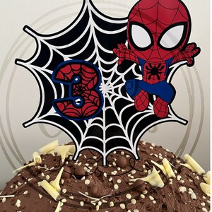 May include: A cake topper featuring a cartoon Spider-Man figure in red and blue, with a black and white spiderweb design and the number 3. The cake is covered in chocolate frosting and decorated with white chocolate shavings.
