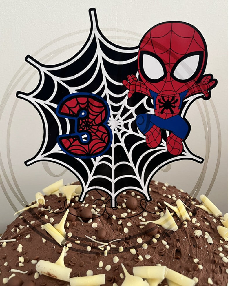May include: A cake topper featuring a cartoon Spider-Man figure in red and blue, with a black and white spiderweb design and the number 3. The cake is covered in chocolate frosting and decorated with white chocolate shavings.