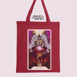 Puede incluir: Bolsa de tela roja con una impresión de lienzo blanco de una carta de tarot que representa a Los Enamorados. La carta está rodeada de un marco dorado y presenta una ilustración colorida de dos figuras con alas.