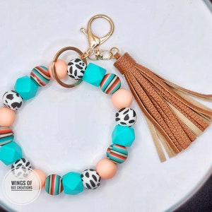May include: A turquoise and peach beaded keychain with a brown tassel. The beads have different patterns, including stripes, animal print, and solid colors. The keychain is attached to a gold key ring.