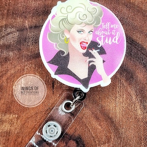 May include: A round, pink retractable badge reel featuring a retro illustration of a woman with blonde hair, red lipstick, and a cigarette. The text "Tell me about it stud" is on the badge. The reel has a clear plastic strap and a metal clip.