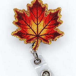 May include: A maple leaf badge reel featuring a gradient of red, orange, and yellow colors with a textured, glittery finish. The leaf is outlined in gold glitter and attached to a clear plastic strap with a metal snap button fastener.