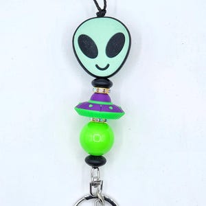 May include: A lanyard with an alien theme. The top features a mint green alien face with black eyes and a smile. Below is a green and purple UFO bead, a green bead, and a black cord. A silver-colored clasp and key ring are at the bottom.