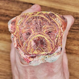 May include: A colorful, abstract pug face design. The artwork features shades of red, yellow, and pink, with a clear, textured border. The image is held against a wooden background.