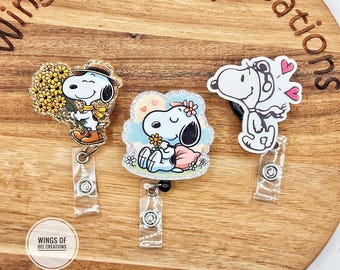 May include: Three Snoopy-themed badge reels. One features Snoopy holding flowers, another shows Snoopy with a flower crown, and the third has Snoopy with hearts. Each reel has a clear plastic retractable cord and a metal clip.