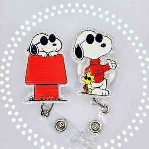 May include: Two Snoopy badge reels. One features Snoopy on his red doghouse, the other shows Snoopy in a red sweater with the text "Joe Cool". Both have black sunglasses and are outlined with silver glitter.