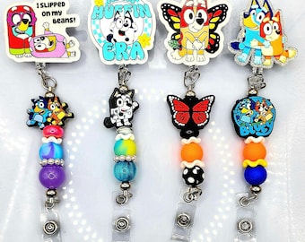 May include: Four retractable badge holders with cartoon character designs. Each holder features a different character, with colorful beads and a clear plastic clip. The designs include characters with text that reads "I slipped on my beans!" and "In my muffin era."