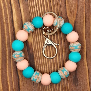 May include: A beaded keychain wristlet featuring a mix of peach, turquoise, and patterned beads. The wristlet has a gold-colored clasp and key ring. The patterned beads have a Southwestern design in shades of orange, teal, and cream.