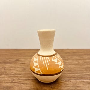 May include: A small, white ceramic vase with a brown and white geometric design. The vase has a round body and a narrow neck.