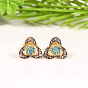 May include: A pair of stud earrings featuring a triangular shape with rounded edges. The centre of each earring contains a cluster of small, bright blue stones surrounded by a gold-toned frame and an outer border of tiny, dark, textured stones.
