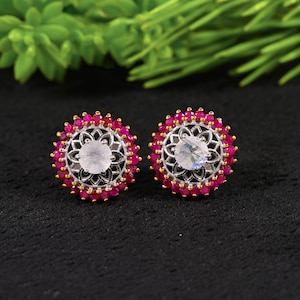 Könnte beinhalten: Ein Paar runde Ohrstecker mit einem zentralen, schillernden Mondstein-Cabochon, umgeben von einem silbernen Filigranrahmen und einem äußeren Rand aus kleinen, leuchtend pinkfarbenen Rubinen, die in goldfarbenen Krappen gefasst sind.