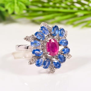Puede incluir: Un anillo de plata con una gran piedra central de rubí ovalada, rodeada de piedras azules en forma de lágrima de zafiro y pequeños diamantes. El anillo tiene un diseño floral, sobre un fondo blanco brillante con follaje verde.