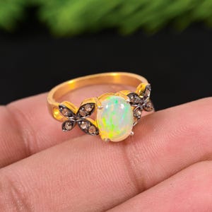 May include: A gold ring featuring an oval opal gemstone with a vibrant play of color. The ring is adorned with butterfly-shaped accents set with small, dark gemstones. The band has a polished finish, creating a striking contrast with the opal.