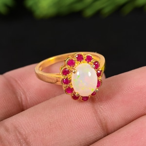 Op de afbeelding: Een gouden ring met een ovale, iriserende opaal als middensteen, omringd door kleine, rode edelstenen. De ring heeft een klassiek, elegant ontwerp met een gouden band.