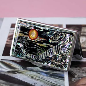 May include: A rectangular metal business card case featuring an iridescent mother-of-pearl inlay on the front. The design depicts a tiger in a landscape with tall grass and a prominent red circular sun against a black background.