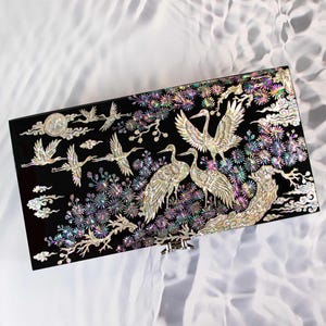 May include: A rectangular black box featuring an intricate mother-of-pearl inlay design of multiple cranes in flight and standing among stylised pine trees. The iridescent shell pieces create a shimmering effect against the dark surface.