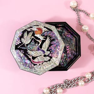 May include: An octagonal black lacquer box with an iridescent mother-of-pearl inlay depicting three cranes, a red sun, and clouds. The lid is partially open, revealing a shimmering abalone shell interior, accompanied by a silver chain with pearls.