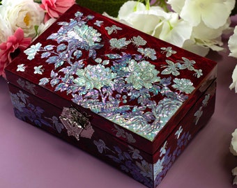 May include: A rectangular, burgundy jewelry box with iridescent floral and butterfly inlay. The box features a silver-toned clasp and is decorated with mother-of-pearl. White and pink flowers are in the background.