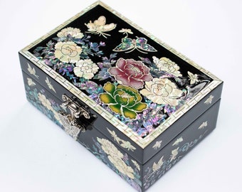 Op de afbeelding: Zwarte rechthoekige sieradendoos met bloemen- en vlindermotieven. De doos heeft inlegwerk van parelmoer in wit, roze, groen en iriserende kleuren. Een zilverkleurige sluiting zekert de doos, die versierd is met bloemen en vlinders.