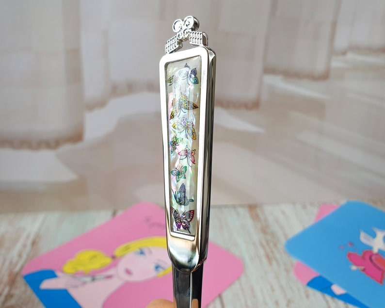 May include: A silver letter opener with a decorative handle featuring a row of colorful butterflies. The handle is made of mother of pearl and has a silver bow at the top.