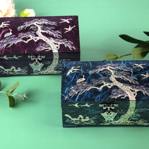 K&ouml;nnte beinhalten: Zwei dekorative Boxen mit aufwendigen Baum- und Vogelmotiven. Die Boxen sind rechteckig mit einem dunkelvioletten und blauen Hintergrund. Der Baum und die V&ouml;gel sind in einer helleren Farbe gehalten, und die Boxen haben einen Schmetterlingsverschluss.